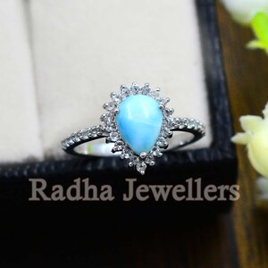 May include: A pearlescent blue gemstone ring with a halo of smaller white gemstones. The ring is set in silver and has a delicate band.