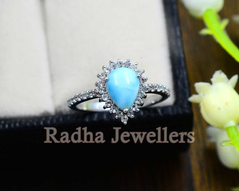 May include: A pearlescent blue gemstone ring with a halo of smaller white gemstones. The ring is set in silver and has a delicate band.