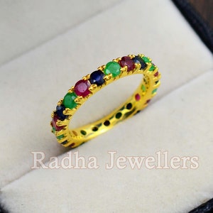 Może przedstawiać: Złoty pierścionek z rzędem kolorowych kamieni szlachetnych. Pierścionek jest ozdobiony zielonymi, czerwonymi i niebieskimi kamieniami.