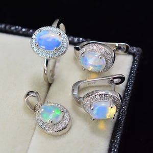 Peut inclure: Un ensemble de bijoux comprenant une bague, un pendentif et des boucles d'oreilles en opale. Chaque pièce présente une opale ovale centrale entourée d'un halo de petits gemmes clairs. Les opales affichent des teintes bleues, vertes et jaunes, serties dans du métal argenté.
