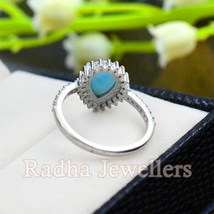 May include: A silver ring with a teardrop-shaped turquoise stone surrounded by smaller clear stones. The ring is in a black ring box with the words "Radha Jewellers" printed on the inside.