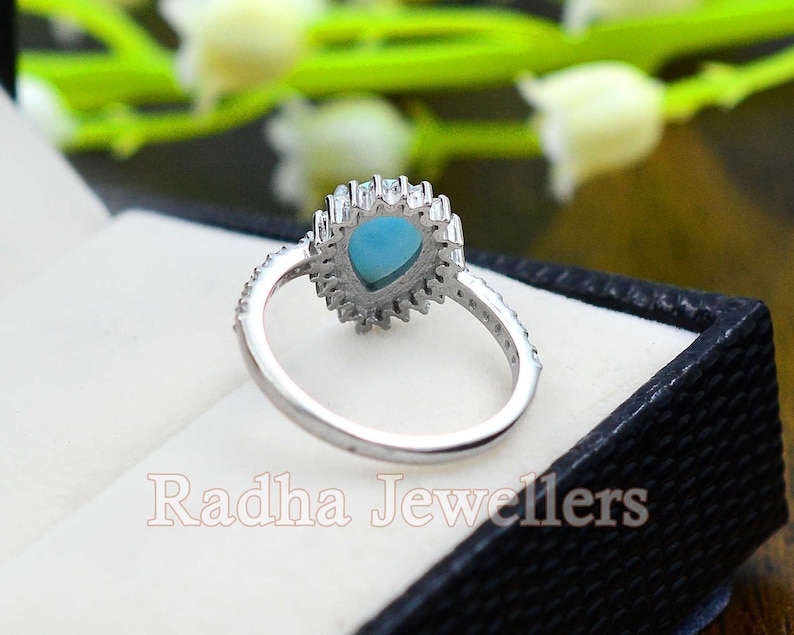 May include: A silver ring with a teardrop-shaped turquoise stone surrounded by smaller clear stones. The ring is in a black ring box with the words "Radha Jewellers" printed on the inside.