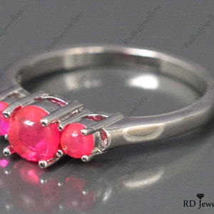 May include: A silver ring with three pink gemstones set in a three-prong setting. The ring is on a gray surface.