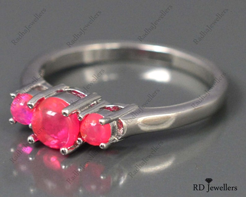 May include: A silver ring with three pink gemstones set in a three-prong setting. The ring is on a gray surface.