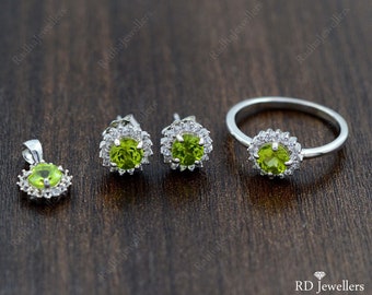 May include: A jewelry set featuring a pendant, stud earrings, and a ring. Each piece has a central, round, light green gemstone surrounded by smaller, clear stones. The metal appears to be silver or white gold. The set is displayed on a dark wooden surface.