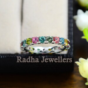 May include: A silver band ring with a row of colorful gemstones. The ring features a variety of colors, including green, blue, pink, and yellow.