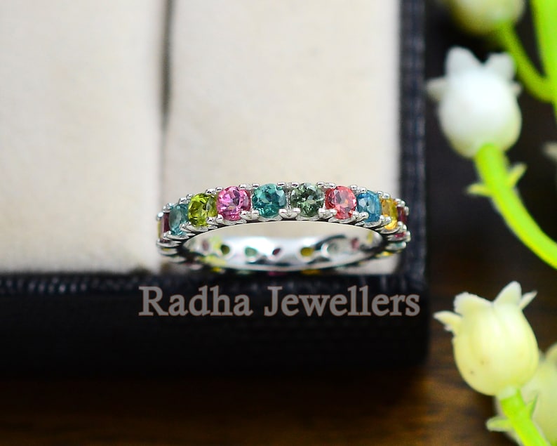 May include: A silver band ring with a row of colorful gemstones. The ring features a variety of colors, including green, blue, pink, and yellow.