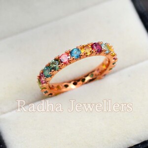 May include: A rose gold band ring with a rainbow of colorful gemstones. The ring is set in a white velvet box with the text "Radha Jewellers" printed on the bottom.