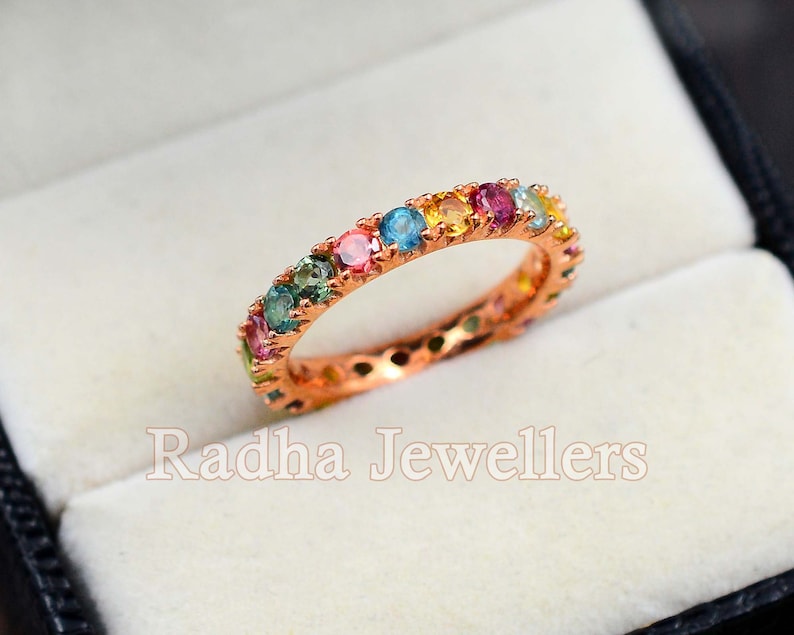 May include: A rose gold band ring with a rainbow of colorful gemstones. The ring is set in a white velvet box with the text "Radha Jewellers" printed on the bottom.