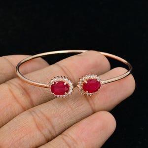 Puede incluir: Un brazalete abierto de tono oro rosa con dos rub&iacute;es de color rojo intenso en talla ovalada, cada uno rodeado por un halo de peque&ntilde;as piedras blancas transparentes. El brazalete tiene una banda de metal fina y lisa que se curva hacia los engastes.
