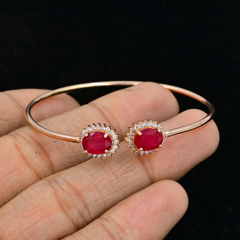 Puede incluir: Un brazalete abierto de tono oro rosa con dos rub&iacute;es de color rojo intenso en talla ovalada, cada uno rodeado por un halo de peque&ntilde;as piedras blancas transparentes. El brazalete tiene una banda de metal fina y lisa que se curva hacia los engastes.