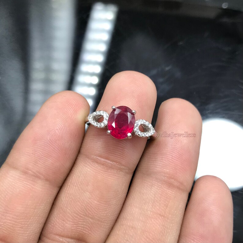 May include: A silver ring with a central oval ruby, complemented by two pear-shaped accents set with smaller, clear stones. The ring is shown on a hand.