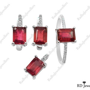 Puede incluir: Un conjunto de joyería compuesto por un anillo, un colgante y un par de pendientes de metal plateado. Cada pieza presenta una piedra roja rectangular de corte esmeralda sujeta por cuatro garras, con pequeñas piedras blancas en los engastes.