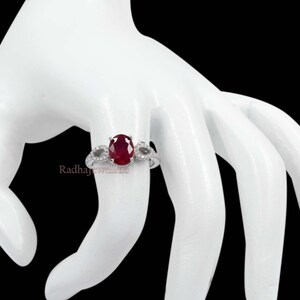 May include: A silver ring featuring a large oval ruby gemstone as the central focus, flanked by two smaller accent stones. The ring is displayed on a white hand against a black backdrop. The text "Radhajewellers" is visible.