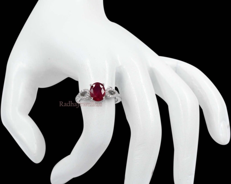 May include: A silver ring featuring a large oval ruby gemstone as the central focus, flanked by two smaller accent stones. The ring is displayed on a white hand against a black backdrop. The text "Radhajewellers" is visible.