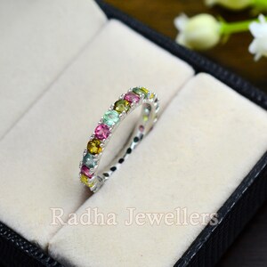 May include: A silver band ring with a row of colorful gemstones. The ring is set in a white velvet box with the text "Radha Jewellers" printed on the bottom.