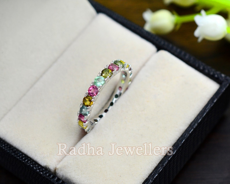 May include: A silver band ring with a row of colorful gemstones. The ring is set in a white velvet box with the text "Radha Jewellers" printed on the bottom.
