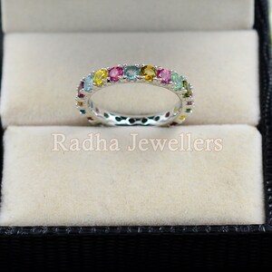 May include: A silver band ring with a colorful row of gemstones. The ring is in a black ring box with a white interior.