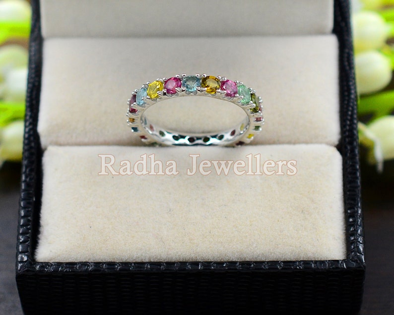 May include: A silver band ring with a colorful row of gemstones. The ring is in a black ring box with a white interior.