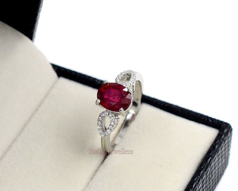 May include: A silver ring featuring a large, oval-cut ruby gemstone. The ring has a delicate design with small, clear accent stones. The ring is displayed in a black jewellery box with a white interior.