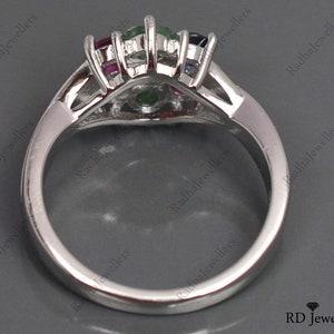 May include: A silver ring featuring a cluster of colourful gemstones, including emerald, ruby, sapphire, and diamonds. The ring is set in a white metal band.