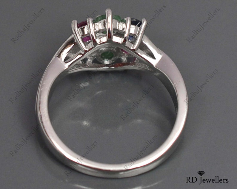 May include: A silver ring featuring a cluster of colourful gemstones, including emerald, ruby, sapphire, and diamonds. The ring is set in a white metal band.