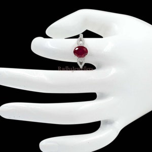 May include: A silver ring with a large oval-cut ruby gemstone. The ruby is a deep red colour and is surrounded by smaller, clear gemstones. The ring has a delicate, openwork design on the band. The text "Radhajewellers" is visible.