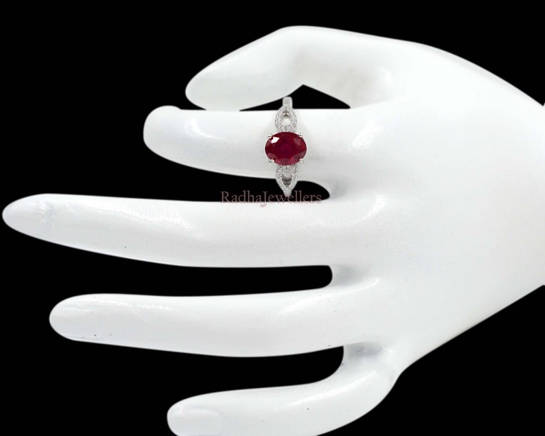 May include: A silver ring with a large oval-cut ruby gemstone. The ruby is a deep red colour and is surrounded by smaller, clear gemstones. The ring has a delicate, openwork design on the band. The text "Radhajewellers" is visible.