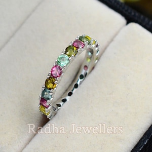 May include: A silver band ring with a row of colorful gemstones. The gemstones are green, pink, yellow, and blue.