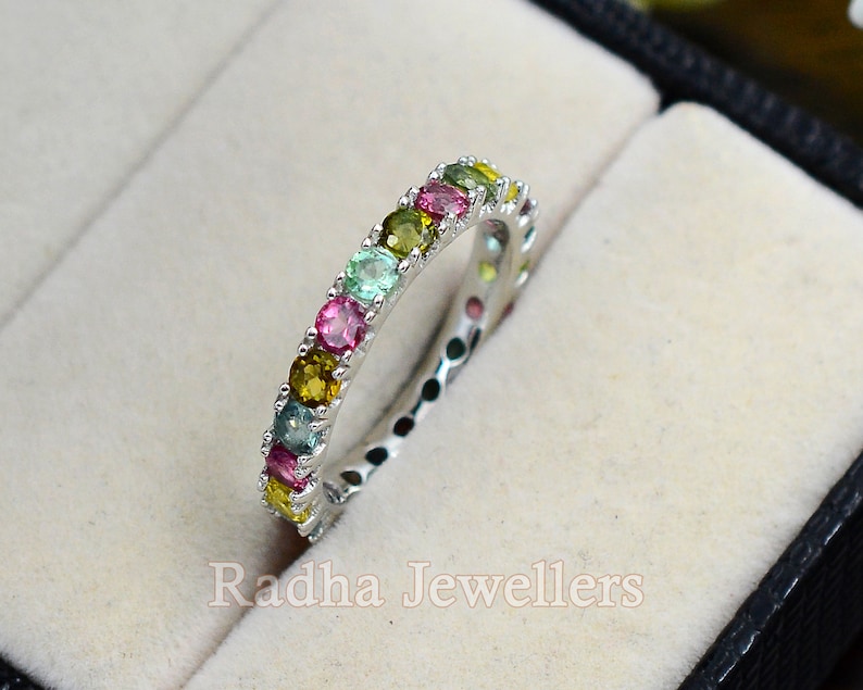 May include: A silver band ring with a row of colorful gemstones. The gemstones are green, pink, yellow, and blue.