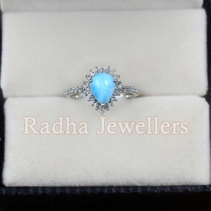 May include: A pear-shaped blue gemstone ring with a halo of smaller white gemstones. The ring is set in silver and is displayed in a black ring box with the text "Radha Jewellers".