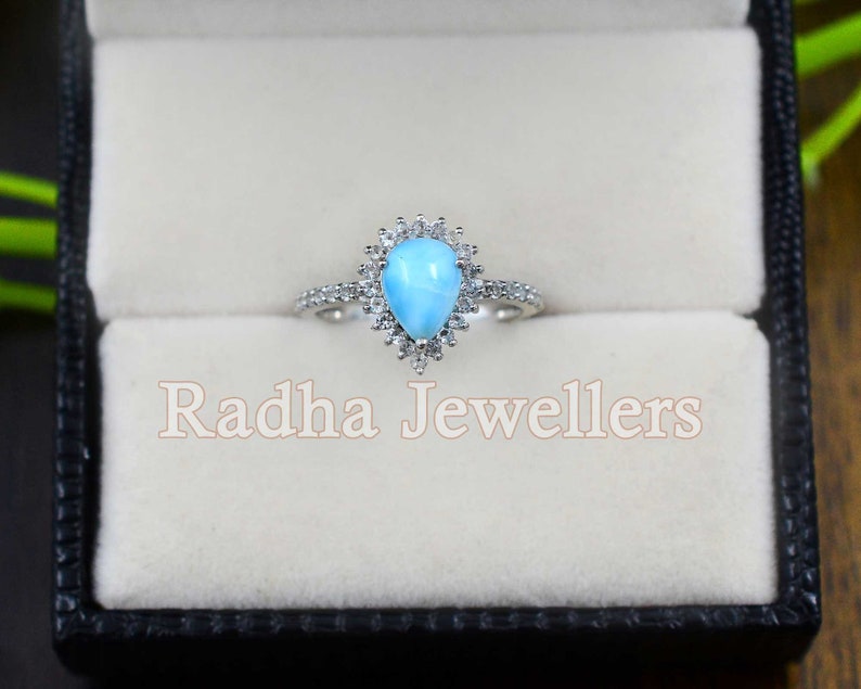 May include: A pear-shaped blue gemstone ring with a halo of smaller white gemstones. The ring is set in silver and is displayed in a black ring box with the text "Radha Jewellers".