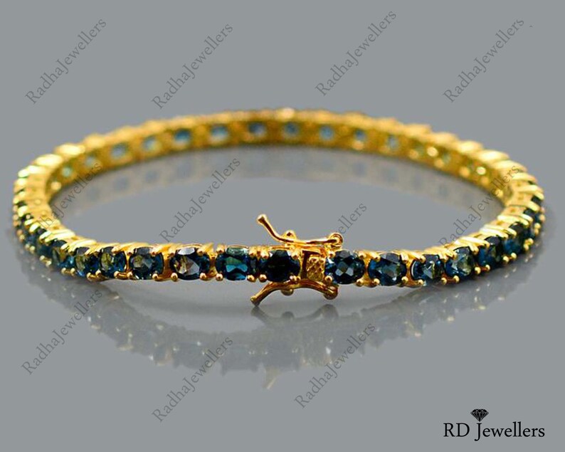 May include: A gold tennis bracelet with a row of blue gemstones. The bracelet has a clasp closure.