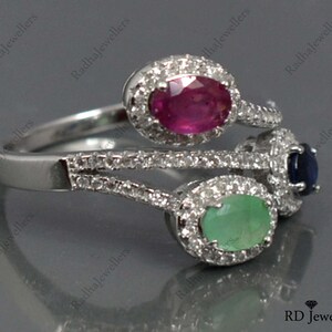 Op de afbeelding: Een zilveren ring met drie ovale edelstenen: een robijn, een smaragd en een saffier. Elke edelsteen is omgeven door een halo van kleine, heldere stenen. De ring heeft een uniek, gelaagd ontwerp.