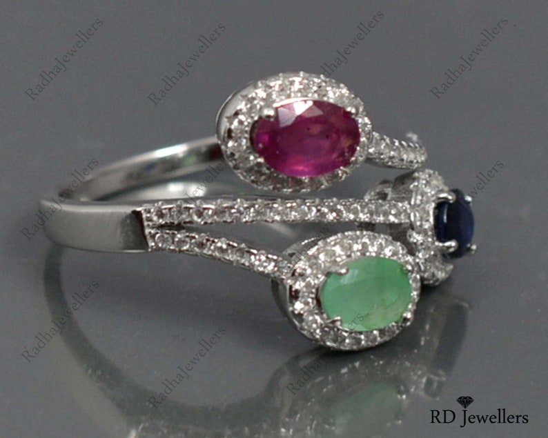 Op de afbeelding: Een zilveren ring met drie ovale edelstenen: een robijn, een smaragd en een saffier. Elke edelsteen is omgeven door een halo van kleine, heldere stenen. De ring heeft een uniek, gelaagd ontwerp.