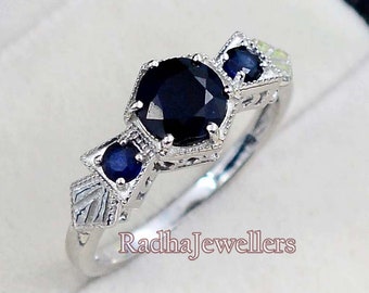 May include: A silver ring featuring a large, round, dark blue gemstone at the center, flanked by two smaller, round, dark blue gemstones. The ring has an intricate, filigree design and the text "RadhaJewellers".