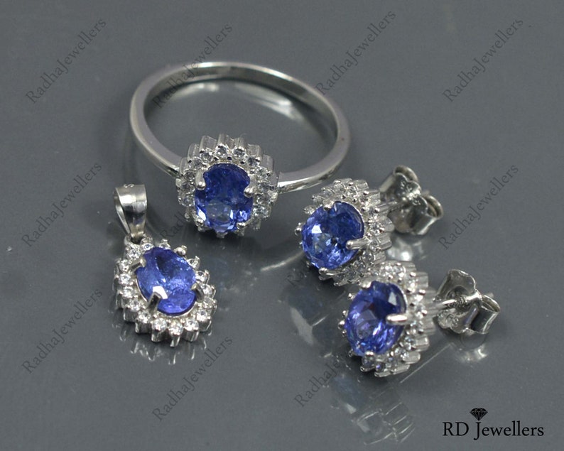 May include: A set of sterling silver jewelry featuring a ring, pendant, and earrings. Each piece has a blue gemstone surrounded by a halo of smaller white gemstones.