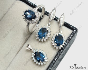 Può includere: Un anello d'argento, un paio di orecchini e un ciondolo, tutti con una pietra ovale blu circondata da pietre bianche più piccole. I gioielli sono esposti in una scatola di velluto nero.