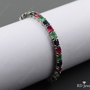 May include: A silver-toned tennis bracelet featuring a repeating pattern of oval-cut red, green, and dark blue gemstones set in a metal frame. The bracelet is displayed wrapped around a white cylindrical form with the text RD Jewellers visible.