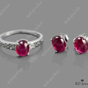 May include: A silver ring and matching stud earrings set. The ring and earrings feature oval-shaped, deep red gemstones. The ring has a decorative band. The earrings are simple studs. The jewellery is displayed on a grey surface.