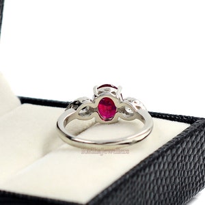 May include: A silver ring featuring a large oval ruby gemstone. The ring has a delicate design with smaller accent stones on either side of the main stone. The ring is presented in a black jewellery box.