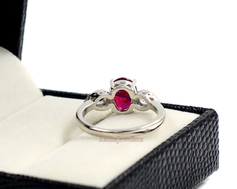 May include: A silver ring featuring a large oval ruby gemstone. The ring has a delicate design with smaller accent stones on either side of the main stone. The ring is presented in a black jewellery box.