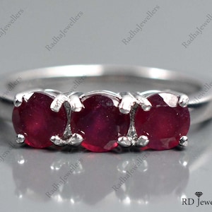 Könnte beinhalten: Ein silberner Ring mit drei roten Rubin-Steinen, die in die Band eingefasst sind. Der Ring liegt auf einer grauen Oberfläche.