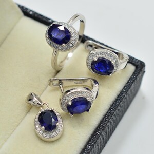 Puede incluir: Un conjunto de joyas que incluye un anillo, pendientes y un colgante. Cada pieza tiene un engaste plateado y una gran gema ovalada de color azul oscuro, rodeada de piedras más pequeñas y claras. El conjunto se exhibe en una caja de joyería negra.