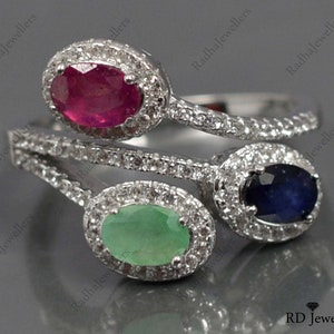 Op de afbeelding: Een zilveren ring met drie ovale edelstenen: een robijn, een smaragd en een saffier. Elke edelsteen is omgeven door een halo van kleine, heldere stenen. De ringband is ook versierd met kleine, heldere stenen.