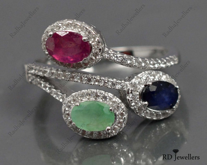 Op de afbeelding: Een zilveren ring met drie ovale edelstenen: een robijn, een smaragd en een saffier. Elke edelsteen is omgeven door een halo van kleine, heldere stenen. De ringband is ook versierd met kleine, heldere stenen.