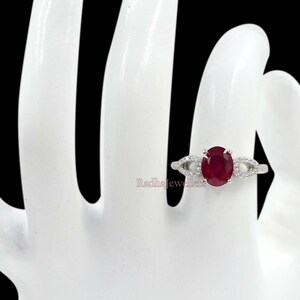 May include: A silver ring with an oval-cut, deep red ruby gemstone. The ring has a delicate design with small, clear accent stones on either side of the ruby. The ring is set on a white hand against a black background.