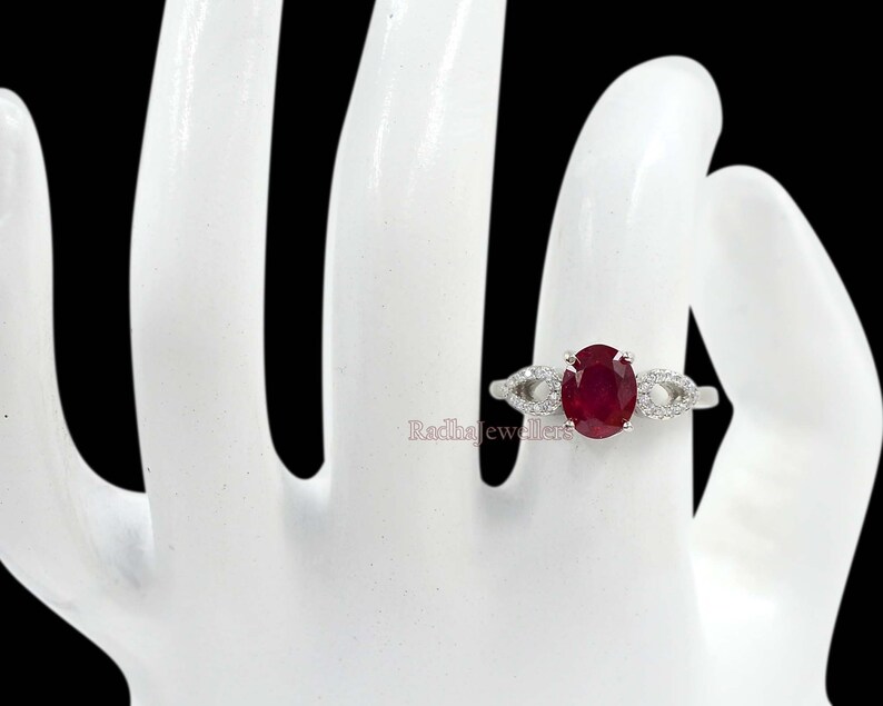 May include: A silver ring with an oval-cut, deep red ruby gemstone. The ring has a delicate design with small, clear accent stones on either side of the ruby. The ring is set on a white hand against a black background.
