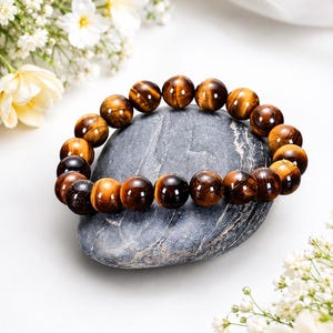 May include: A beaded bracelet featuring polished, spherical tiger's eye gemstones. The beads display a natural chatoyant luster with alternating bands of golden-brown and dark brown colors, strung together in a continuous circular loop.