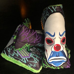 May include: A black and green golf club head cover with a white, blue, and red Joker face design. The cover has a green fuzzy lining.